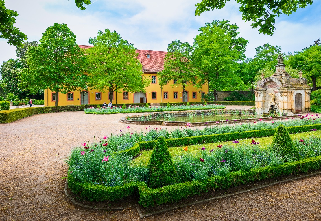 Oßmannstedt-Wielandgut_Matthias Eckert_WLT2023 (23) Der Park ist mit gepflegten weiten Kieswegen, mit von kleinen Hecken umgrenzten Blumenbeten und einer Brunnenanlage mit großem Wasserbecken ausgestattet. Im Hintergrund das Herrenhaus mit gelber Fassade und rotem Ziegeldach.